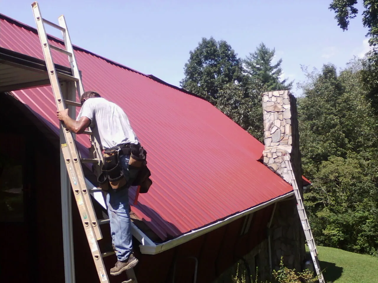 Roofer on a ladder beside a red metal roof and stone chimney — Soffit & Fascia Repair by Roofing Brothers in Carolina
